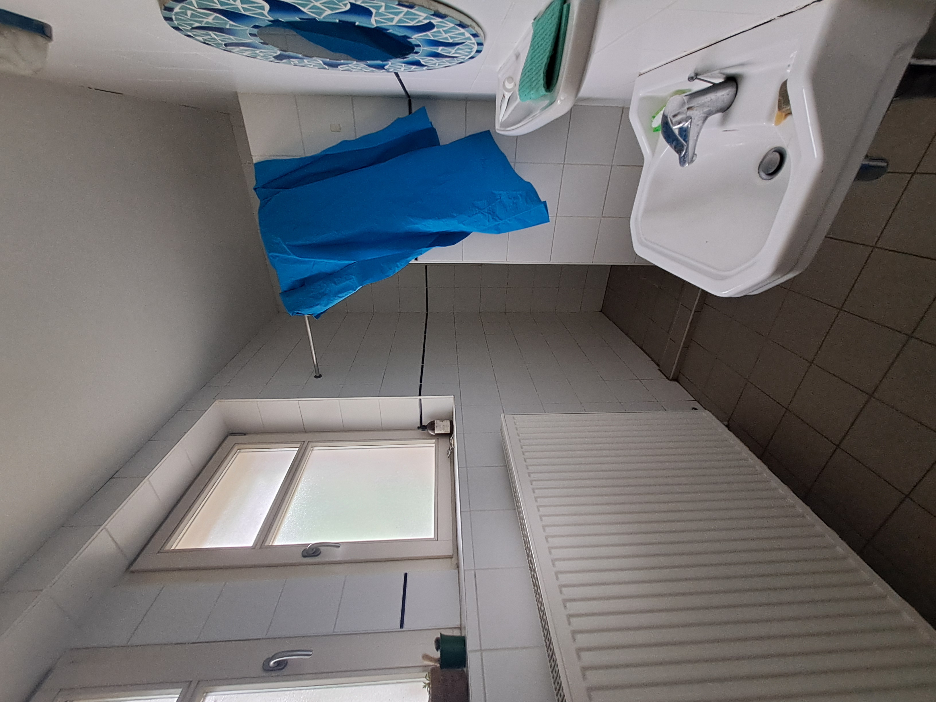 une salle de bain avec une radiateur et deux fenetres à gauche un lavabo à droite et une douche à rideau au fond